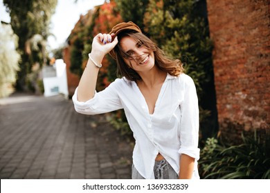 Wind Flinches Wavy Hair Of Young Woman Wearing Corduroy Hat. Shot Of Girl In White Blouse And Gray Shorts On Old Street With Bushes