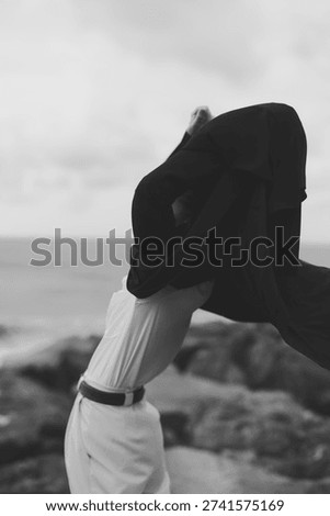 Wind flows through a scarf draped over the head, white outfit on a rocky coast, creating a dramatic, monochrome silhouette, and seashells on shore, mood of resilience and solitude