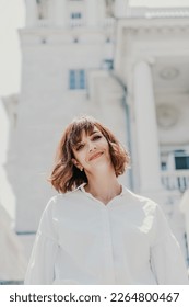 Wind hair style. A portrait of a woman outdoors, her shoulder-length brown hair blowing in the wind. Dressed in a white shirt against a light building. - Shutterstock ID 2264800467