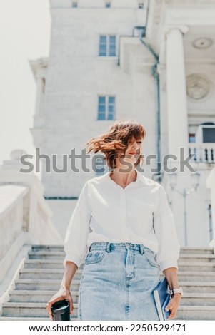 Wind hair style. Portrait of a woman outdoors, her shoulder-length brown hair blowing in the wind. Dressed in a white shirt and denim skirt against a light building.