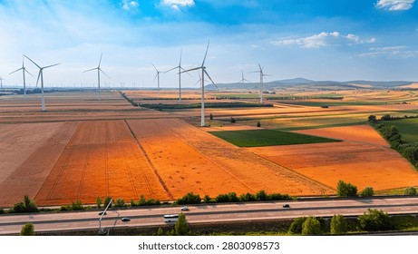 Wind turbines stand in open fields. Wind turbines turn in the breeze over large fields during the day. Cars travel on the road beside the fields.