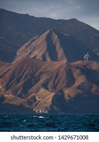 Wind Turbines In The Top Of A Volcanic Mountain Cliff At Sunset. Eolian Farm, Renewable Energy In Front A Climate Change. View From  The Sea And Shore On The Island Of Cape Verde.