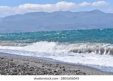 Wind-driven waves sweep toward the beach, backed by hazy mountains and scattered clouds on the horizon.