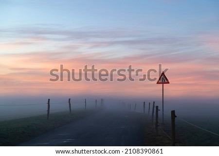 Winding country road with signpost “bottleneck“ in Iserlohn Sauerland Germany on a misty foggy winters evening. Course of Street with fence and delineators vanishing in wafts of mist on the horizon.