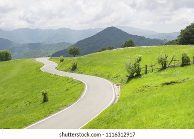 Winding Curvy Rural Road In Green Meadows And Beautiful Mountain Views In Countryside In Natural Spring