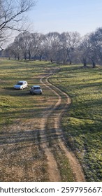 A winding dirt path meanders through an open field, flanked by leafless trees under a clear sky. Two parked cars rest on the grass, adding a human element to the tranquil rural scene.