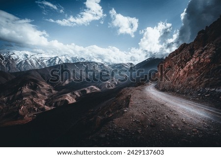 A winding dirt road cuts through the rugged terrain of the Atlas Mountains, edged precariously by a steep cliff. The snow-capped peaks in the distance stand in stark contrast to the rocky foreground. 