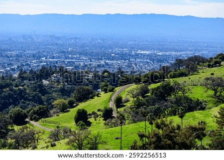 A winding dirt trail bordered by fencing cuts through green hills with scattered trees, overlooking Downtown San Jose skyline and the surrounding suburban sprawl from Alum Rock Park