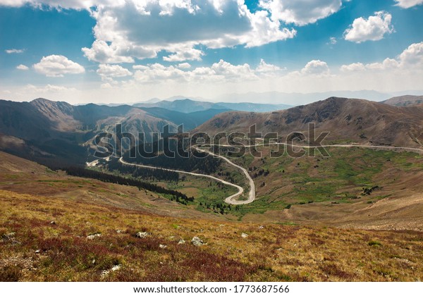 Winding meandering road through the mountains at\
the continental divide on US highway 6 in Colorado. Orange and\
green grasses in the landscape with tall mountain peaks. Vacation\
destination panorama