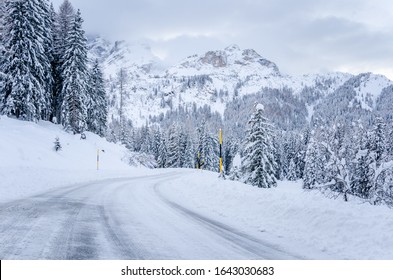 Winding mountain road covered in fresh snow at the foot of towering mountains on a cloudy winter day. Concept of inclement weather and dangerous driving conditions.