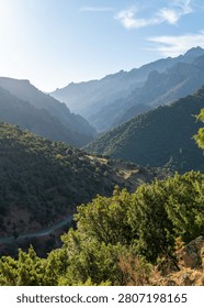 Winding mountain road threads through a sun-drenched valley, framed by hazy, layered peaks in the distance.