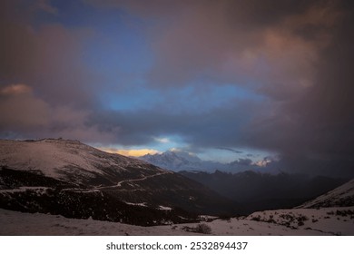 A winding mountain road under a dramatic sky with moody clouds, distant snowy peaks, and a serene, dusky atmosphere.