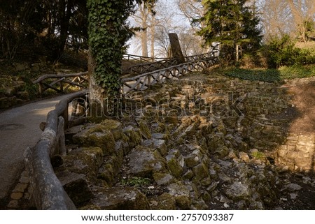 A winding park path bordered by rustic wooden railings and moss covered rocks in warm natural light. Peaceful outdoor landscape with strong leading lines, textured stone surfaces, and a calm woodland atmosphere, walking, and nature themes. 