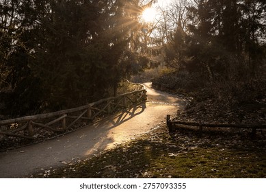 A winding park path lined with rustic wooden fencing and lit by warm low sunlight filtering through tall trees. Atmospheric outdoor scene with soft shadows, natural textures, and a peaceful woodland mood, suitable for travel, and relaxation concepts.
