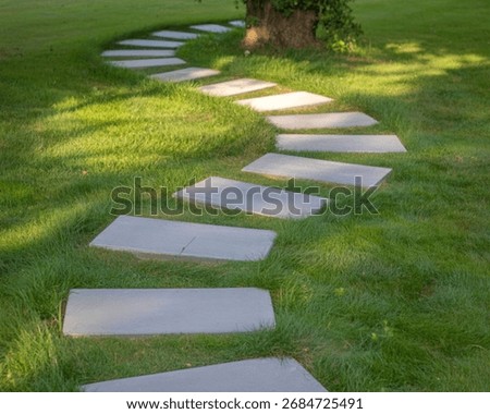 A winding path of flat, grey stone pavers is laid across a vibrant green lawn. Sunlight dapples the grass, creating a serene and inviting garden scene.