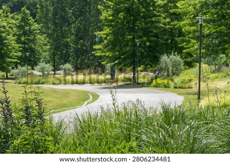 A winding paved path passes through lush trees and ornamental plants. The landscaped park offers a peaceful outdoor setting.