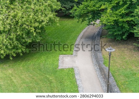 A winding paved path passes through a quiet park bordered by mature trees and grass. The peaceful route encourages walking and relaxation in nature.