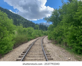 A winding railroad track cuts through the dense, green forests of the Utah mountains, framed by towering peaks and a dramatic, cloud-filled sky. - Powered by Shutterstock