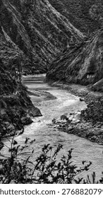 A winding river cuts through a dramatic canyon, with sharp cliffs and textured slopes on both sides. In black and white, the contrast highlights the rugged terrain and flowing water, while a narrow path and distant building emphasize isolation and depth.