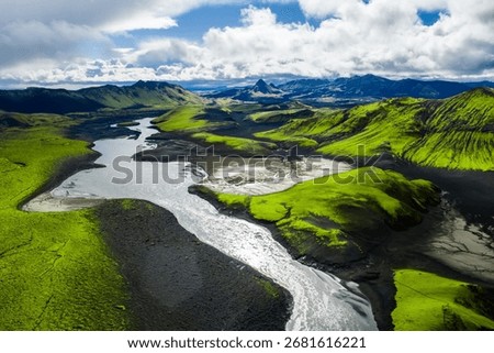 A winding river flows through a valley of green moss covered hills and dark volcanic soil, with jagged peaks under a partly cloudy sky in Iceland.