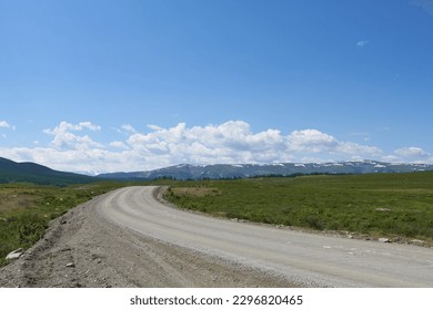 A winding road against a blue sky with clouds and mountain peaks on the horizon. A steep turn to the right.