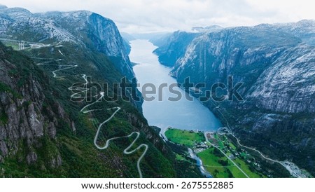 A winding road carves through the dramatic cliffs of Lysefjord, leading down to the serene waters below. Lush greenery contrasts against the rugged mountains in this stunning Norwegian setting.