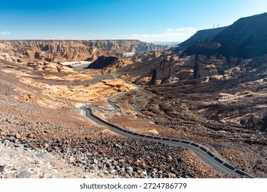 A winding road cuts through a dramatic desert canyon in Saudi Arabia, with layered red rocks, rugged hills, and a distant horizon under bright daylight. Ideal for travel, nature, and scenery photography.