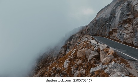 A winding road cuts through a foggy, rocky mountainous terrain, with mist partially obscuring the mountain peak, creating a mysterious and serene atmosphere. 🌫️🗻 - Powered by Shutterstock