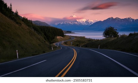 A winding road leads through lush hills toward distant snow-capped peaks, with a serene lake and a pastel sky creating a tranquil atmosphere. - Powered by Shutterstock