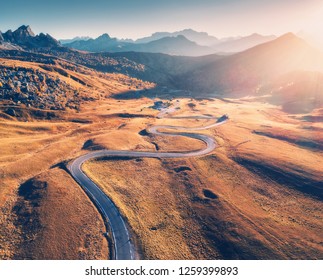 Winding Road In Mountain Valley At Sunset In Autumn. Aerial View Of Asphalt Road In Passo Giau. Dolomites, Italy. Top View Of Roadway, Mountains, Meadows With Orange Grass, Blue Sky And Gold Sunlight