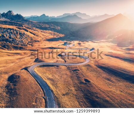 Winding road in mountain valley at sunset in autumn. Aerial view of asphalt road in Passo Giau. Dolomites, Italy. Top view of roadway, mountains, meadows with orange grass, blue sky and gold sunlight