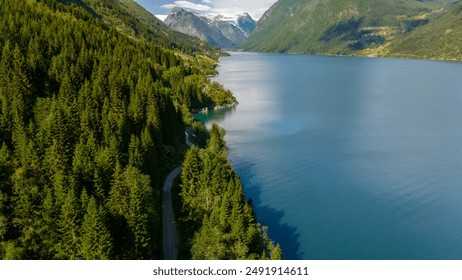 A winding road snakes through a dense forest of evergreen trees, leading to a sparkling blue lake nestled between towering mountains. Fjaerlandsfjorden, Fjord, Vestland, Norway - Powered by Shutterstock