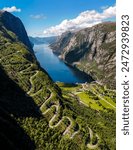 winding road snaking its way up a steep mountainside overlooking a serene fjord in Norway. The road is flanked by lush green vegetation and towering cliffs. Kjerag, Lysebotn, Lysefjorden, Norway