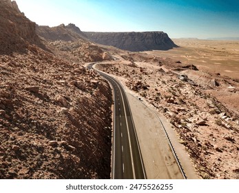 a winding road through Antelope Canyon Pass, flanked by rugged red rock formations and vast desert plains under a clear blue sky. The barren, rocky terrain contrasts with the smooth, dark asphalt. - Powered by Shutterstock