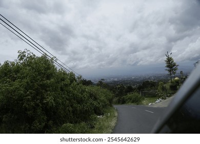 A winding road through a rural area with lush green vegetation on either side. In the distance, a city skyline can be seen through a hazy atmosphere. The sky is overcast with dark clouds, suggesting a - Powered by Shutterstock