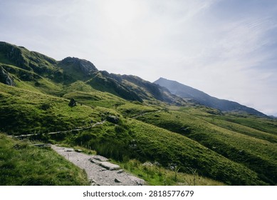 A winding rocky path ascends through vibrant green, grass-covered mountain hills under a bright, hazy sky, leading towards distant peaks in a vast, serene natural landscape.