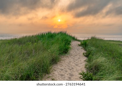 A winding sandy path cuts through lush green dune grass, leading the eye toward a vast, calm ocean. Above the horizon, a glowing sun breaks through a heavy layer of golden and grey clouds at sunset.
