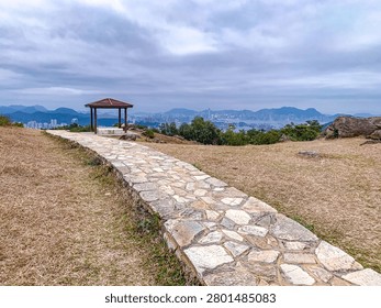 A winding stone path guides visitors towards a wooden gazebo situated on a grassy hilltop with a panoramic view of a sprawling urban landscape and busy port