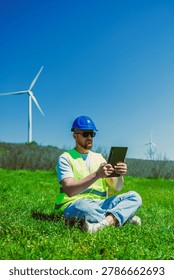 A windmill engineer sits on the grass in a field. He wears a safety vest and hard hat while using a tablet to check equipment near wind turbines during sunny weather.