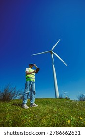 A windmill engineer stands on grass looking at a tablet while inspecting the wind turbine. The sky is clear and blue, and wind turbines are visible in the background.