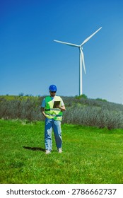 A windmill engineer stands in an open field with green grass, observing data on a tablet near a large wind turbine under a clear blue sky.