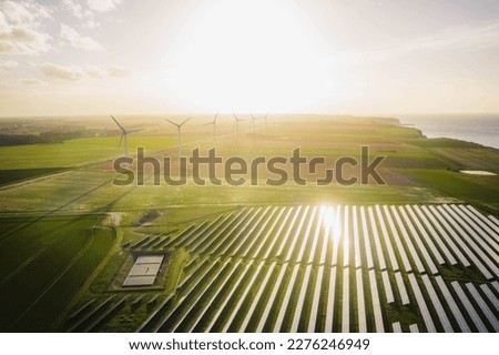 Windmills and solar panels farm in green fields close to the ocean. Renewable energies concept. Green energy for carbon dioxide emission reduction. 