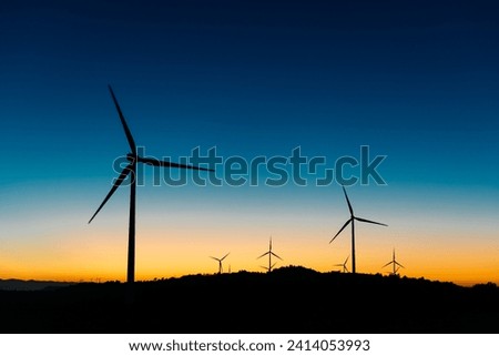 Windmills with wind turbines farm against sundown sky over dark hills in evening time