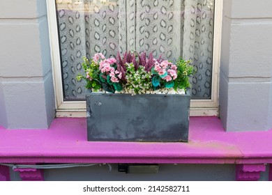 Window box of colourful artificial flowers on a pink window sill with a window with net curtains behind