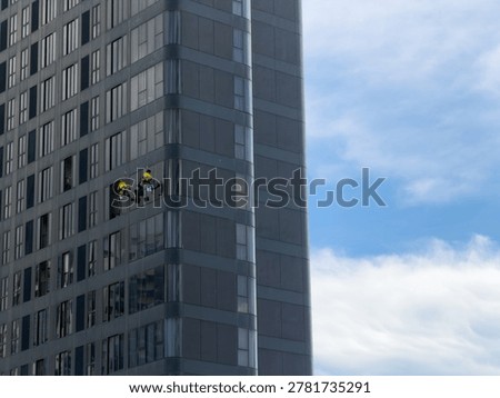 Window cleaners working on a skyscraper glass facade, skyscraper maintenance concept under a blue cloudy sky.