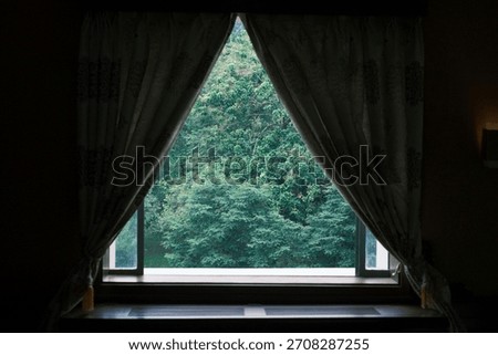 A window with open curtains frames a lush green forest view outside.