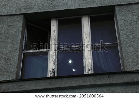 A window with peeling paint and a rickety window on the facade of the students' dormitory. A chandelier and curtains are visible in the window.