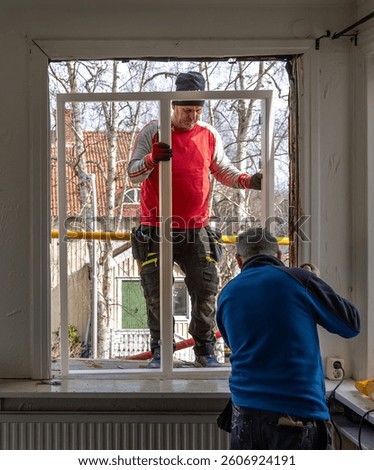 Window replacement underway in a private home. Two tradesmen collaborate to install a new window frame, using hand tools and scaffolding for access and support.