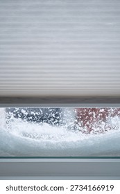 A window with the roller blinds half closed with snow piled against the windows on a winter day.