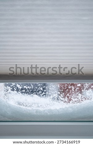 A window with the roller blinds half closed with snow piled against the windows on a winter day.